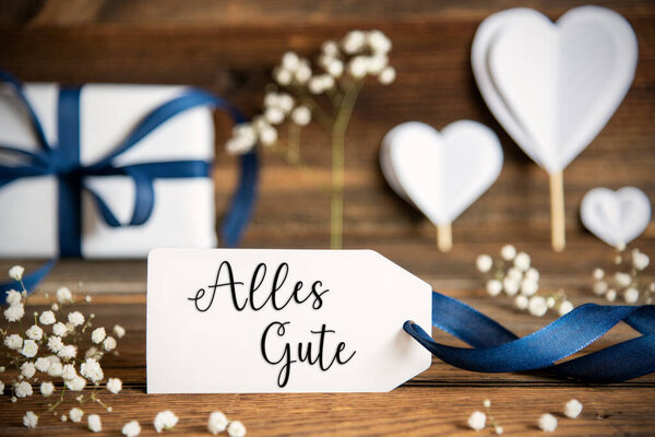 Label With German Text Alles Gute Means Best Wishes. White Festive Decoration Like Present With Blue Bow, Heart and Flowers. Vintage Wooden Background.
