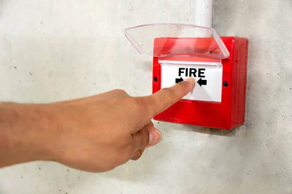 Close up of male hand pointing at red fire alarm switch on concrete wall in office building. Industrial fire warning system equipment for emergency.