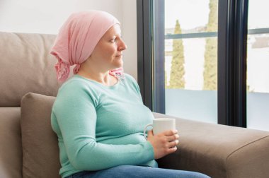 Young woman with cancer in head scarf relaxing drinking coffee sitting on a sofa in the living room in a house interior