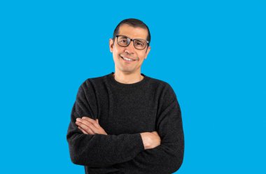 Studio shot of a man wearing casual sweater and glasses over blue background happy face smiling with crossed arms looking at the camera. Positive person.