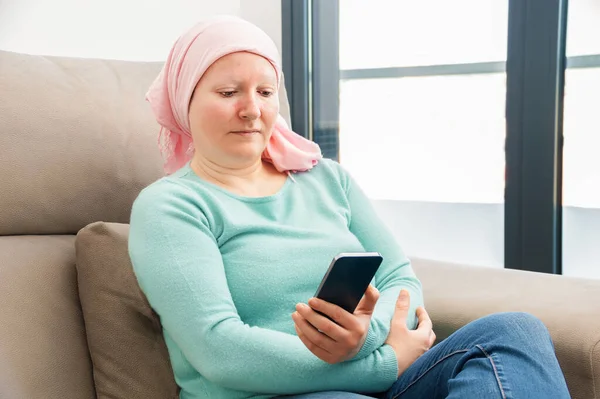 Young woman with cancer in head scarf reading a cancer information in a smart phone sitting on a couch at home