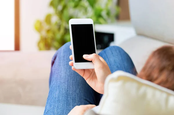 Woman using and showing a blank phone screen lying on a couch at home