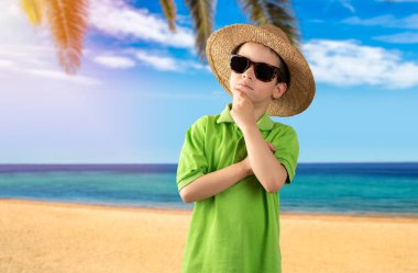 Tropik plajda güneş gözlüğü takan, elleri çenesinde yeşil t-shirt giyen soru soran, düşünceli bir çocuk. Dalgın bir yüz. Şüpheye kapılma ve yana bakma