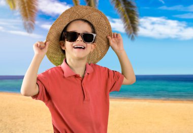 Kırmızı tişörtlü, güneş gözlüklü, tropik plajda gülümseyen ve kameraya bakan bir çocuk.. 