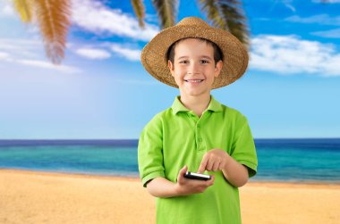 Akıllı telefon kullanan çocuk, mutlu yüzlü ve şapkalı kameraya bakıyor. Kendine güvenen bir gülümsemeyle, tropik plaj arka planında dişlerini gösteriyor.