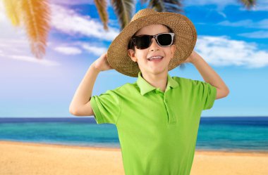 Yeşil tişört giyen, plajda güneş gözlüğü takan, gülümseyen ve kameraya bakan bir çocuk. Pozitif kişi