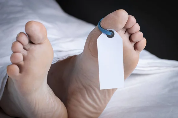 Close-up of the feet of a corpse covered in a sheet; with a blank tag tied on the big toe of the left foot; with an added vignette and copy space