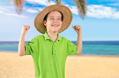 Yeşil tişörtlü tatilci çocuk tropik plajda mutlu ve heyecanlı bir şekilde kolları havada, gülümseyen ve başarı için bağıran bir jest yapıyor..