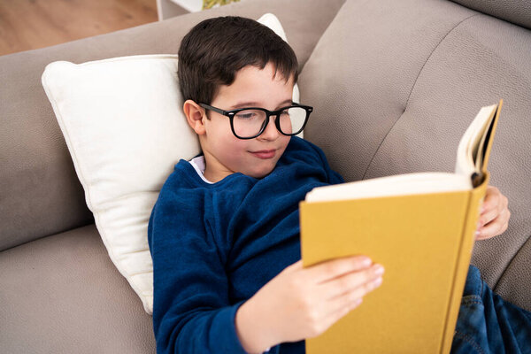 Portrait of cute boy wearing big glasses reading book while lying on couch, homeschooling concept and back to school and copy space