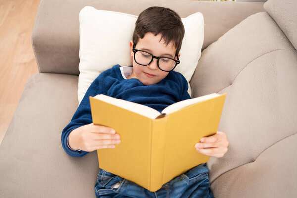 Above view portrait of cute boy wearing big glasses reading book while lying on couch, homeschooling concept and back to school and copy space