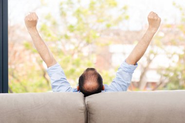 Kel ve kaygısız bir adamın arka görüntüsü. Kolları yukarda bir kanepede ve yeşil arka planda oturma odasının penceresinden dışarı bakıyor.