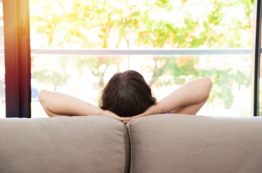 Bekar bir kadının arka görüntüsü pencereden dışarı bakarken evdeki oturma odasındaki kanepede güneş ışığı altında oturuyor.