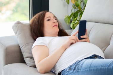 Hamile bir kadının elinde cep telefonuyla kanepede yatarken çekilmiş fotoğrafı.