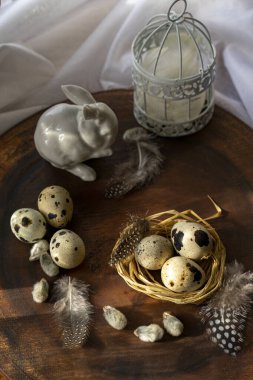 Beyaz porselenden yapılmış bir Paskalya tavşanı ve tüylü bir kafesin arka planında, koyu ahşap bir yüzeyde duran bıldırcın yumurtaları.