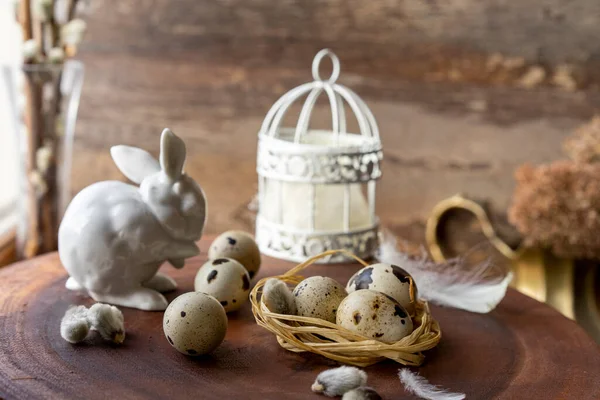 Beyaz porselenden yapılmış bir Paskalya tavşanı ve tüylü bir kafesin arka planında, koyu ahşap bir yüzeyde duran bıldırcın yumurtaları.