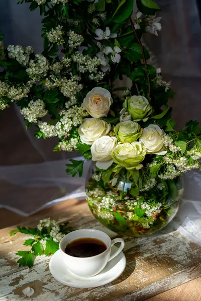 Kırsal ahşap masanın üzerinde bir fincan sade kahve ve güneş ışığı altında cam vazoda bahar beyaz çiçekleri.