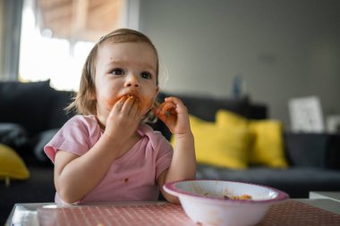 one girl small caucasian toddler female child daughter eating alone at the table at home while watching tv making mess on her face dirty sauce childhood growing up development concept copy space