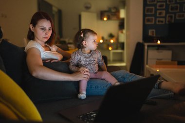 Small caucasian child little girl toddler mother and daughter sitting in dark room at night at home on sofa bed watching cartoons or movies on laptop computer real people family growing up copy space