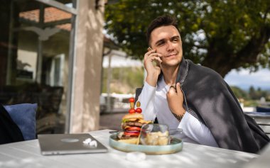 Genç, yetişkin, beyaz bir erkek restoranın terasındaki masada oturuyor. Elinde hamburgerle cep telefonuyla ciddi bir konuşma yapıyor.