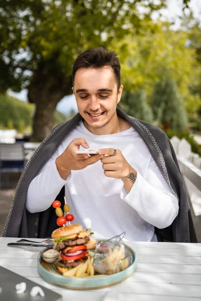 1人の男白人男性がレストランのテラスに座って携帯電話で屋外のテーブルの上の皿の上に食べ物バーガーの写真を撮る本物の人々は幸せな笑顔のスペースをコピー — ストック写真