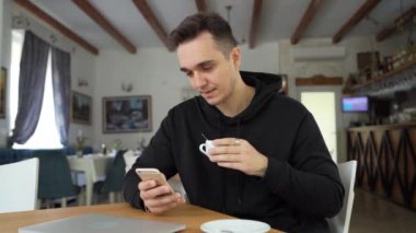 Kafe veya restoranda masada oturan bir adam sıradan giyiniyor cep telefonuyla iletişim kuruyor ve elinde bir fincan gerçek insan kahvesi tutarken konuşuyor.