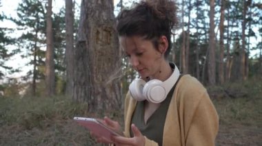 Genç yetişkin bir kadın, parkta ya da ormanda yalnız başına meditasyona hazırlanan kulaklıkları ayarlıyor. Kişisel bakım, zihinsel denge kavramını uyguluyor.