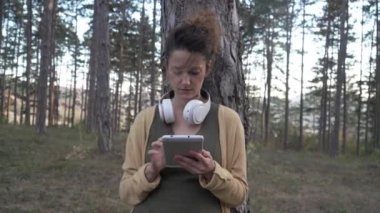 Genç yetişkin bir kadın, parkta ya da ormanda yalnız başına meditasyona hazırlanan kulaklıkları ayarlıyor. Kişisel bakım, zihinsel denge kavramını uyguluyor.