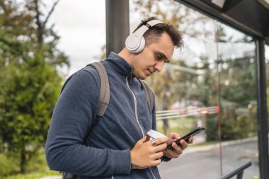Yetişkin bir erkek, kış ya da sonbahar günü sırt çantası öğrencisi ya da turist hayatı fotokopisi çekmek için kulaklık ve cep telefonuyla otobüs durağında bekliyor.