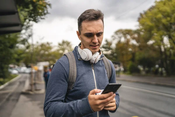 市内の屋外に立つ1人の若い大人の白人男性駅で公共バスの輸送を待っている現代の観光客や学生日に携帯電話のテキストメッセージ本物の人々コピースペース — ストック写真
