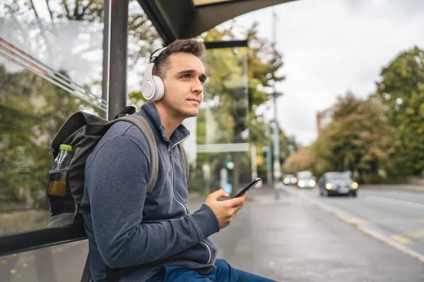 stock image One man young adult male sit at public transport bus station waiting with headphones and mobile smart phone in winter or autumn day with backpack student or tourist city life copy space