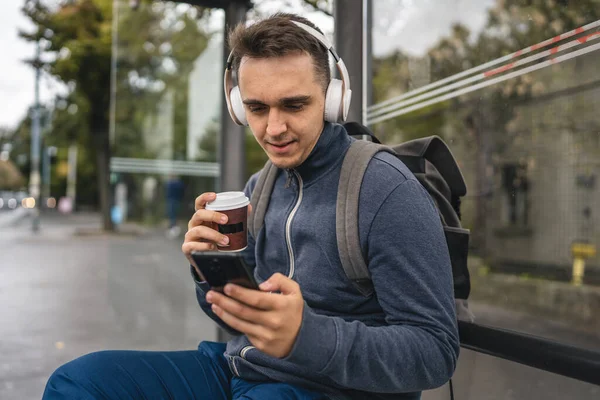 1人の若い大人の男性は 冬または秋の日にヘッドフォンや携帯電話で待っている公共交通機関のバスステーションに座ってバックパックの学生や観光都市の生活コピースペース — ストック写真