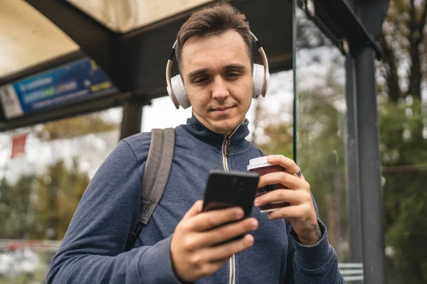 冬または秋の日にヘッドフォンや携帯電話で待っている公共交通機関のバスステーションで1人の若い大人の男性スタンドバックパック学生や観光都市生活コピースペース — ストック写真