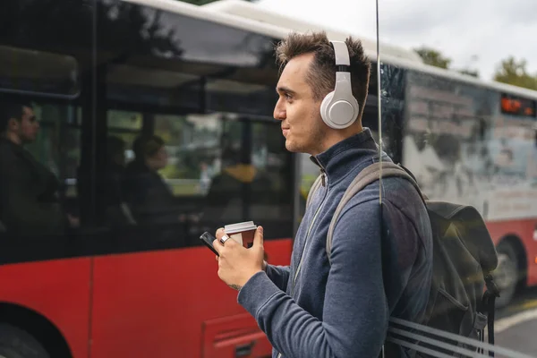 冬または秋の日にヘッドフォンや携帯電話でバスに乗る公共交通機関のバスステーションで1人の若い大人の男性スタンドバックパック学生や観光都市の生活コピースペース — ストック写真