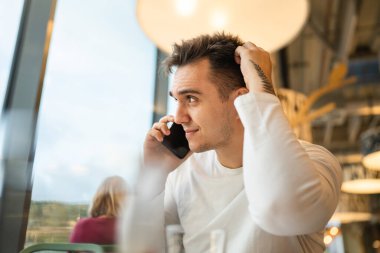 Yetişkin bir erkek, kafeteryada ya da restoranda cep telefonuyla konuşuyor. Gerçek insanlar, düşünceli kafa çözme problemini kopyalıyor.