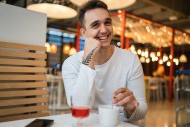 Genç, yetişkin, beyaz bir adam kafeteryada ya da restoranda tek başına oturuyor. Yan tarafa bakıyor, bir fincan kahve içiyor. Gerçek insanlar uzay gülüşünü kopyalıyor.