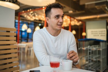 Genç, yetişkin, beyaz bir adam kafeteryada ya da restoranda tek başına oturuyor. Yan tarafa bakıyor, bir fincan kahve içiyor. Gerçek insanlar uzay gülüşünü kopyalıyor.