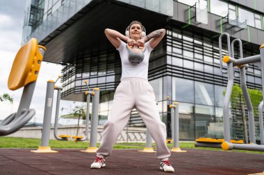 Bir kadın, modern olgun, beyaz kadın. Kısa saçlı, bina önünde günün ilerleyen saatlerinde spor salonunda kettlebell ağırlığı ile spor salonu sağlıklı yaşam tarzı konseptini taklit ediyor.
