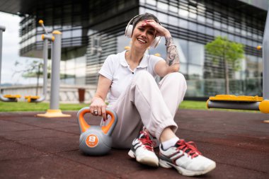 Modern olgun, kısa saçlı, beyaz bir kadın açık hava spor salonunda kettlebell ağırlığıyla oturmuş spor antrenmanı yaptıktan sonra fren yapıyor sağlıklı yaşam tarzı konsepti fotokopi çekiyor.