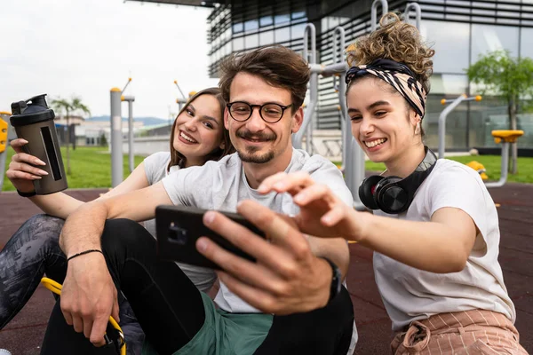 一组男女现代朋友在露天健身房接受训练前或训练后 坐在室外健身公园里 用手机复制自己的照片 — 图库照片