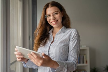 Genç bir kadın, beyaz kadın, iş yerinin penceresinde ya da ön penceresinde dikiliyor. Fotokopi çekiliyor. Fotokopi odası gömleği, kendine güvenen gözlüklü, dijital tablet gülümseyen.