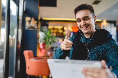 Yetişkin bir erkek Kafe 'de tek başına oturup dijital tablet ve kulaklık kullanıyor. Kulaklıkla çalışıyor ya da uzaktan konferans fotokopisi çekiyor.