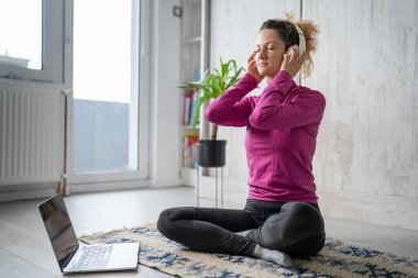Yetişkin bir beyaz kadın milenyum kulaklığı ve dizüstü bilgisayarı kullanarak online meditasyon için bilinçlilik yogası yapıyor. Evdeki gözler kapalı gerçek insanlar mekanı kopyalıyor.