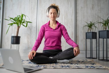 Yetişkin bir beyaz kadın milenyum kulaklığı ve dizüstü bilgisayarı kullanarak online meditasyon için bilinçlilik yogası yapıyor. Evdeki gözler kapalı gerçek insanlar mekanı kopyalıyor.
