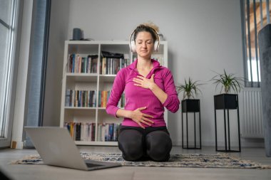 Yetişkin bir beyaz kadın milenyum kulaklığı ve dizüstü bilgisayarı kullanarak online meditasyon için bilinçlilik yogası yapıyor. Evdeki gözler kapalı gerçek insanlar mekanı kopyalıyor.