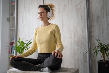 Yetişkin bir beyaz kadın milenyum kulaklıklarını internet rehberli meditasyon için kullanıyor evde gözleri açık zihin yogası yapıyor gerçek insanlar uzay özbakım kavramını taklit ediyorlar.