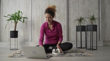 Yetişkin bir beyaz kadın milenyum kulaklığı ve dizüstü bilgisayarı kullanarak online meditasyon yapan evde gözleri kapalı yoga yapan gerçek insanlar.