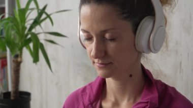 Yetişkin bir beyaz kadın milenyum kulaklıklarını internet rehberli meditasyon için kullanıyor. Evde gözleri kapalı, farkındalık yogası yapıyor. Gerçek insanlar kendi kendilerine bakıyorlar.