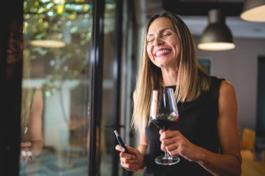 One woman adult middle age caucasian female standing by the window at home or restaurant alone holding glass of red wine and electronic cigarette device happy smile real people copy space