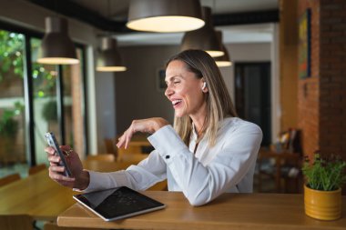 Yetişkin bir orta yaşlı beyaz kadın iş yerinde mutlu bir gülümseme takınıyor, beyaz gömlek giyiyor, ön plan fotoğrafını cep telefonuyla çekiyor ya da kendine güvenen mesajlar yolluyor.