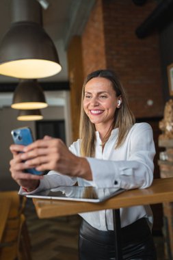 Yetişkin bir orta yaşlı beyaz kadın iş yerinde mutlu bir gülümseme takınıyor, beyaz gömlek giyiyor, ön plan fotoğrafını cep telefonuyla çekiyor ya da kendine güvenen mesajlar yolluyor.